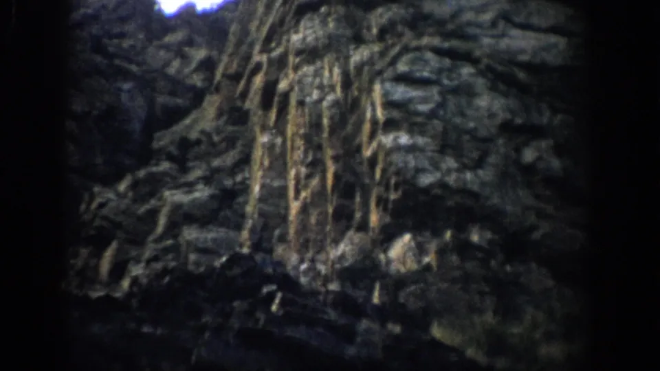 1959 Colorado 8mm frame showing a shadowed rock face with eroded ledges, vertical streaks, and dark viewfinder edges.