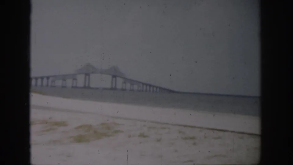 Soft 8mm view of a long bridge extending across water beyond a Florida beach, with faded color and frame wear.