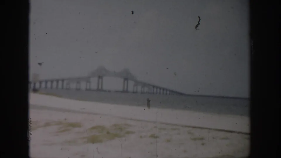 Florida 1959 home-movie frame of a long bridge beyond beach grass and sand, with soft focus and visible film artifacts.