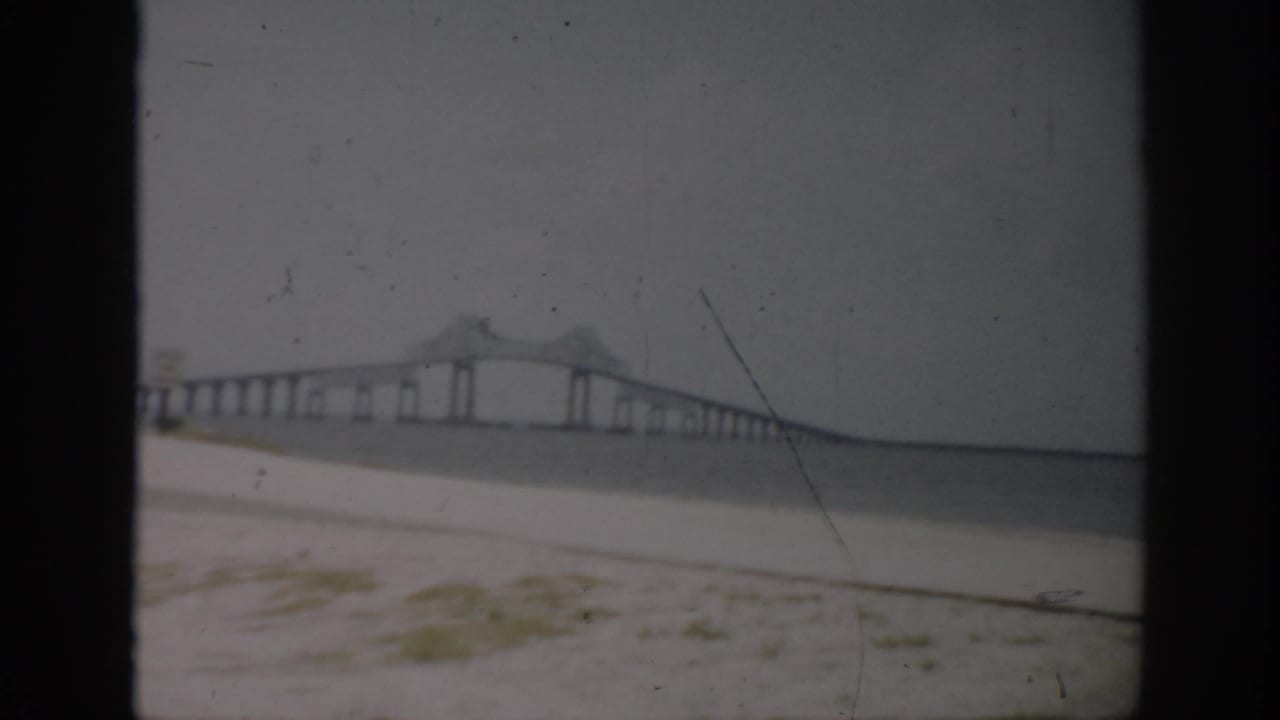 Long bridge viewed from a sandy shoreline across calm water under a gray sky.