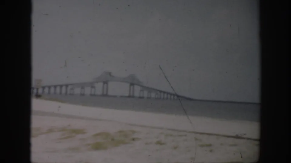 1959 Florida 8mm frame showing a long bridge with repeated piers and a raised central span seen from a pale sandy shore.