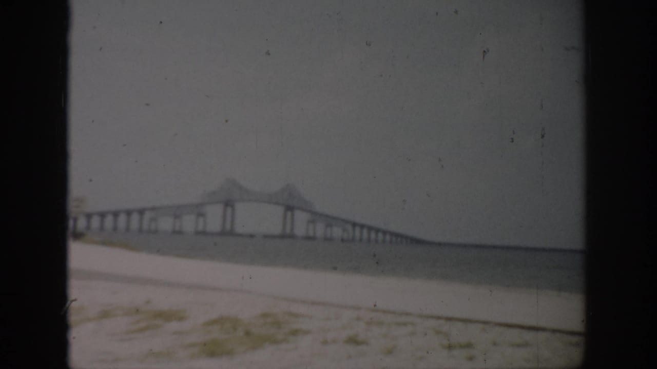 Wide shoreline view of a long bridge extending across water with a central arched section.