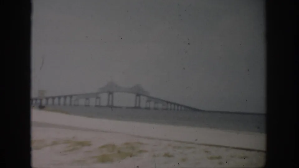 1959 Florida home-movie frame with a distant bridge, repeated piers, calm water, and sandy foreground vegetation.
