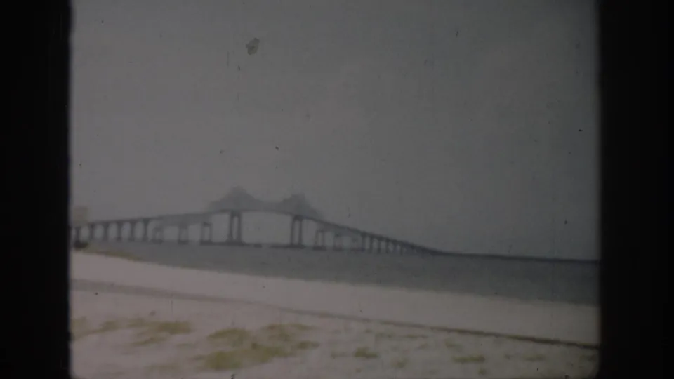 Faded 8mm Florida shoreline view showing a long bridge, calm water, pale sand, and visible dust and scratches.