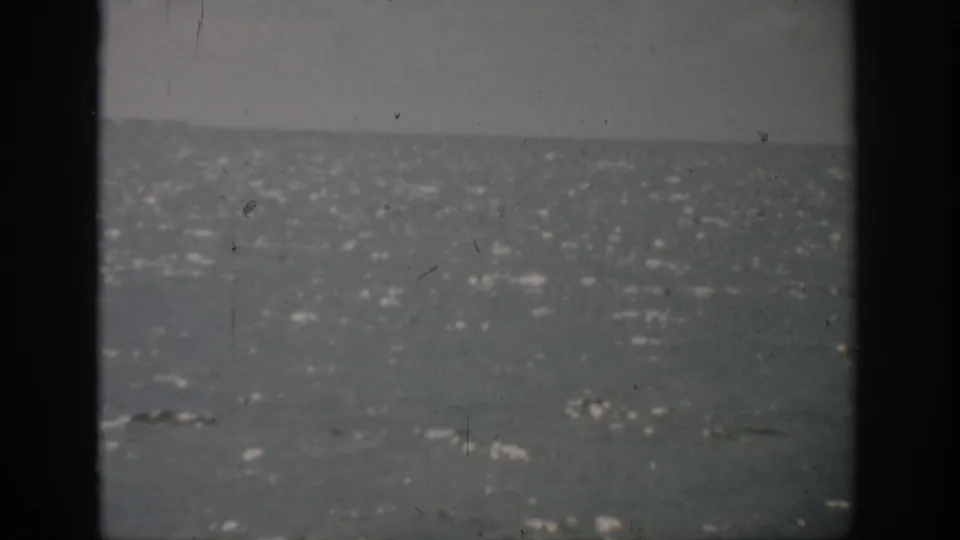 1959 Florida home-movie still with open water, a hazy horizon, gray sky, dark frame edges, and visible 8mm dust and grain.