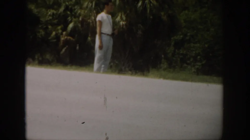 1959 Florida roadside frame showing a man in a white shirt and light pants standing near dense green foliage.