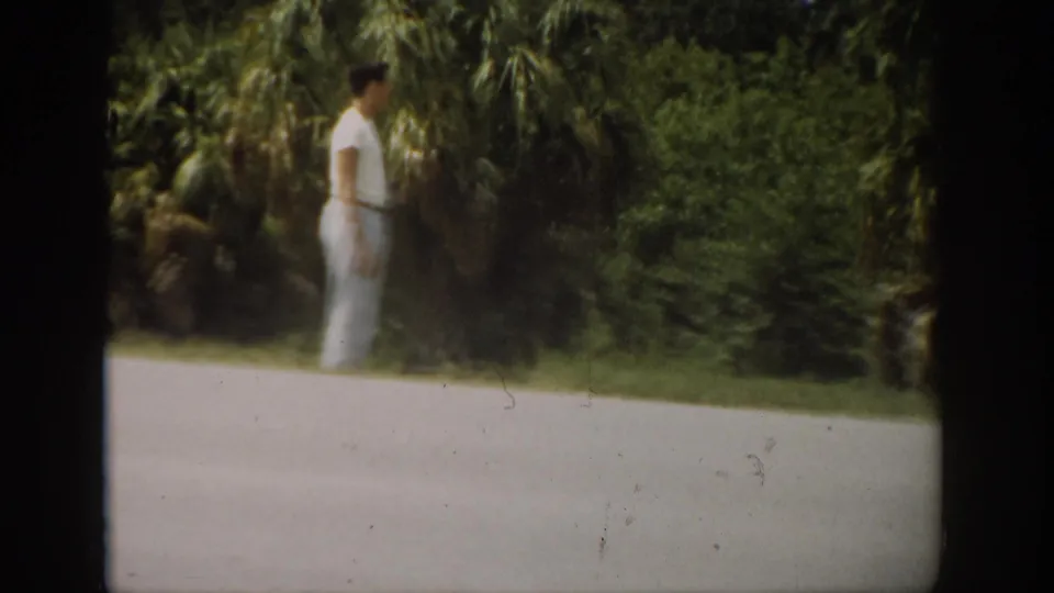 In 1959 Florida home-movie footage, a young man in a white shirt and light pants stands near a paved roadside lined with palms.