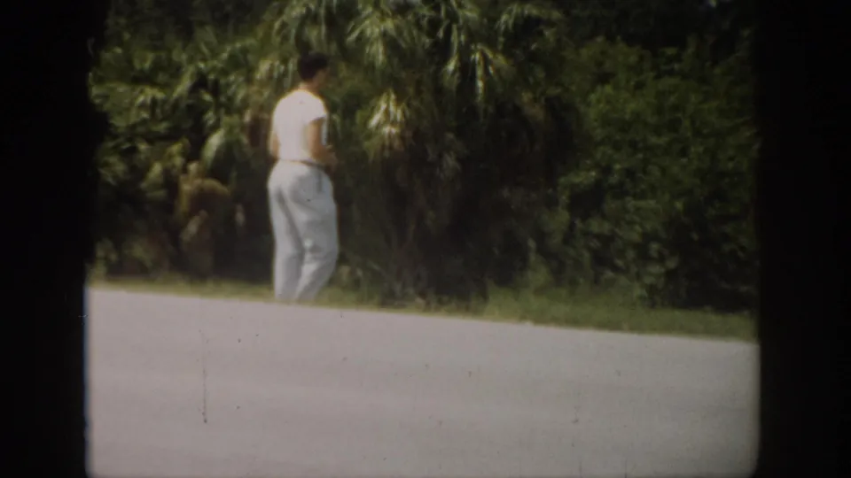 The man turns away from the camera beside the road, with dense palms and roadside grass visible in the 1959 Florida setting.