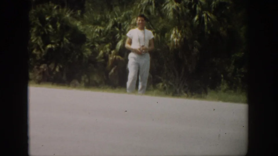 A young man faces the camera in soft 8mm color, standing by a paved Florida roadside with palm trees and foliage behind him.