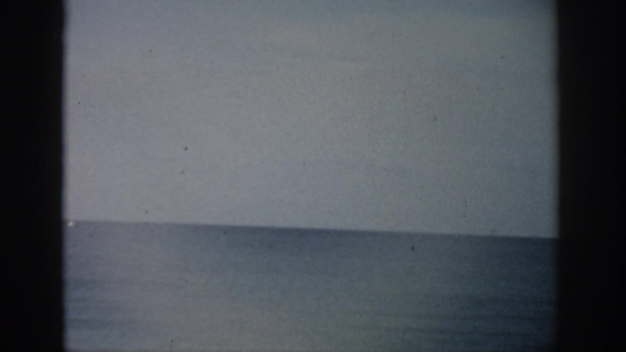 Calm ocean horizon beneath a pale sky with visible film grain and dark frame edges.