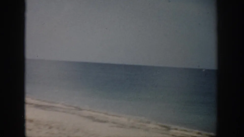 1959 Florida home-movie frame showing a sandy beach edge, gentle surf, calm ocean, and visible 8mm gate border.