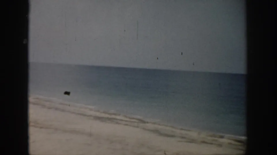 1959 Florida 8mm frame of a calm beach horizon, pale sand, soft surf, and a small dark object near the shore.