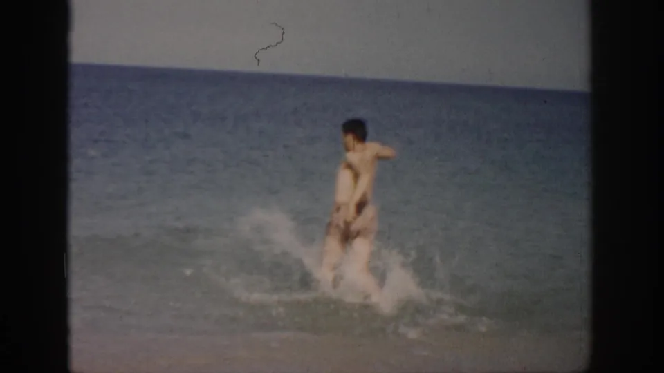 1959 Florida home-movie frame showing a man in patterned swim trunks running through shallow surf toward the ocean.