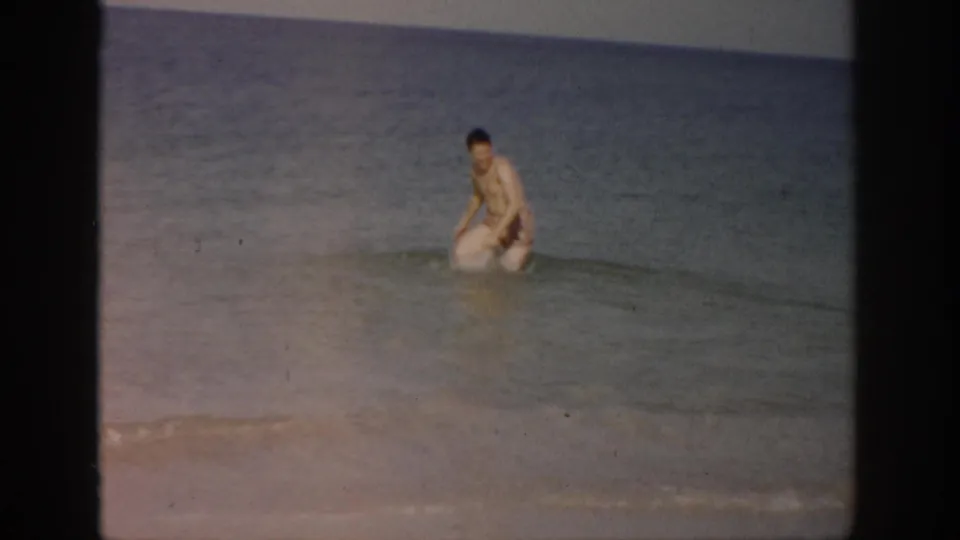The man lowers himself into the shallow Florida surf, with wet sand and soft 8mm color visible around him.