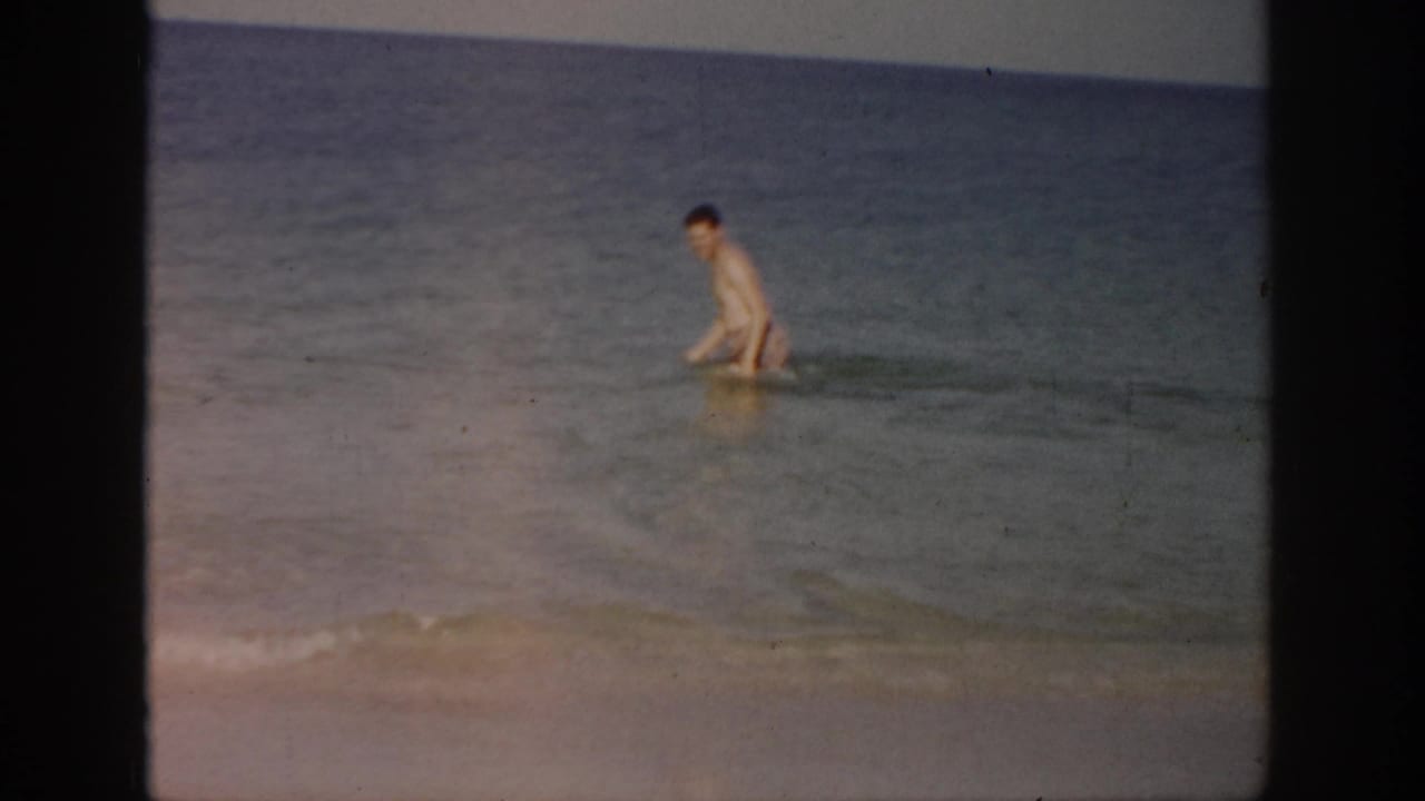 1959 Florida home-movie frame showing a man wading in shallow ocean surf with the horizon behind him.