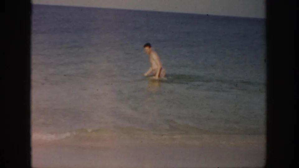 1959 Florida home-movie frame showing a man wading in shallow ocean surf with the horizon behind him.