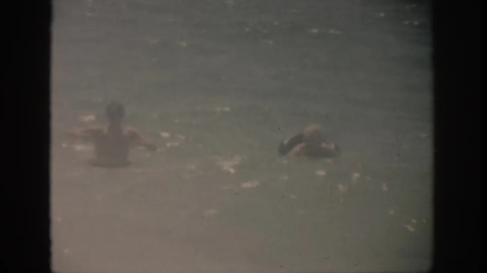 1959 Florida home-movie frame showing swimmers low in the water amid pale surf and soft film grain.