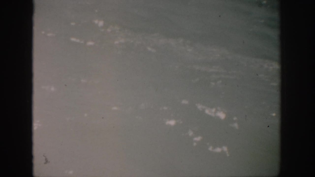 1959 Florida 8mm frame showing hazy blue-green water, small white foam patches, dust specks, and dark gate edges.