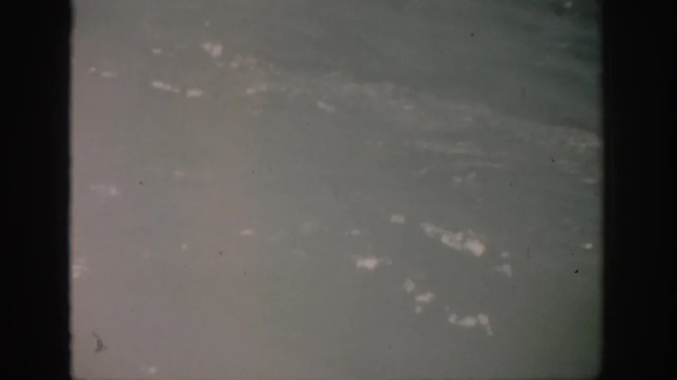 1959 Florida 8mm frame showing hazy blue-green water, small white foam patches, dust specks, and dark gate edges.