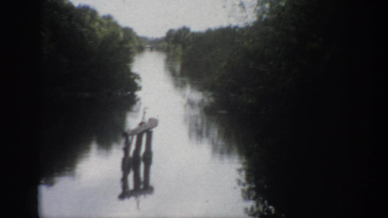 1959 Florida 8mm frame showing a calm river, dense tree-lined banks, and a small wooden post structure reflected in the water.