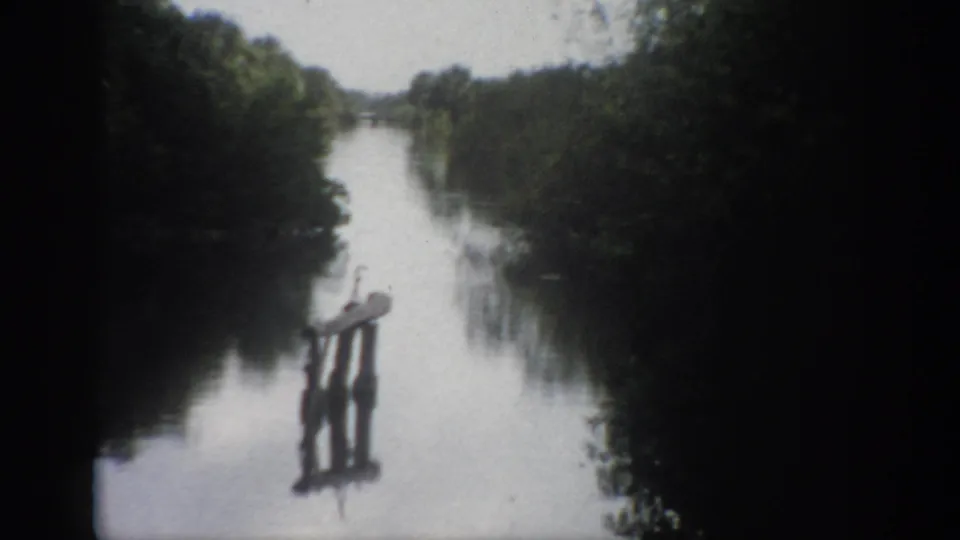 1959 Florida 8mm frame showing a calm river, dense tree-lined banks, and a small wooden post structure reflected in the water.