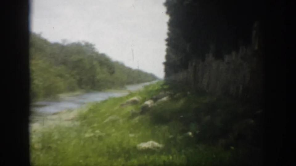 1959 Florida travel footage frame with a winding river, bright green grass, and forested bank seen through dark vehicle framing.
