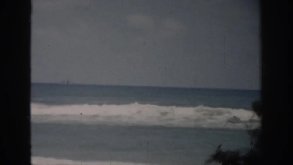 1959 Florida coastal home-movie frame with pale ocean waves, flat horizon, and dark vertical borders suggesting a view through a frame or window.
