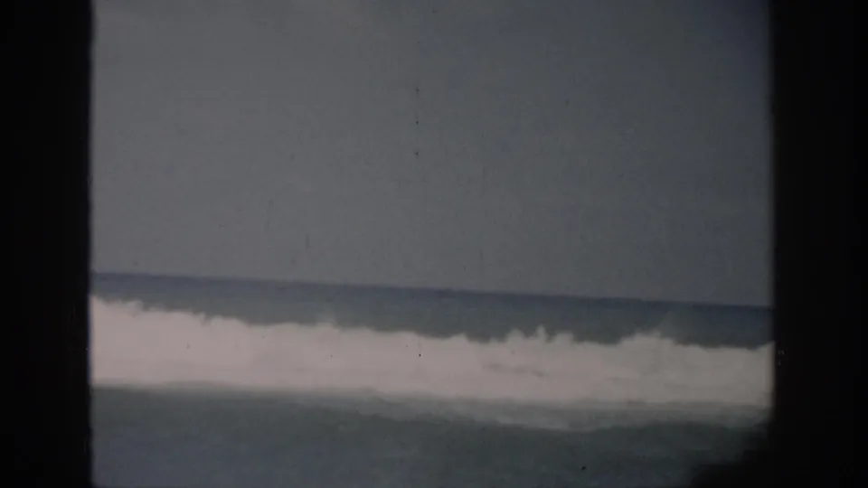 1959 Florida 8mm shoreline view with white surf bands, muted water, and dark foreground frame edges.