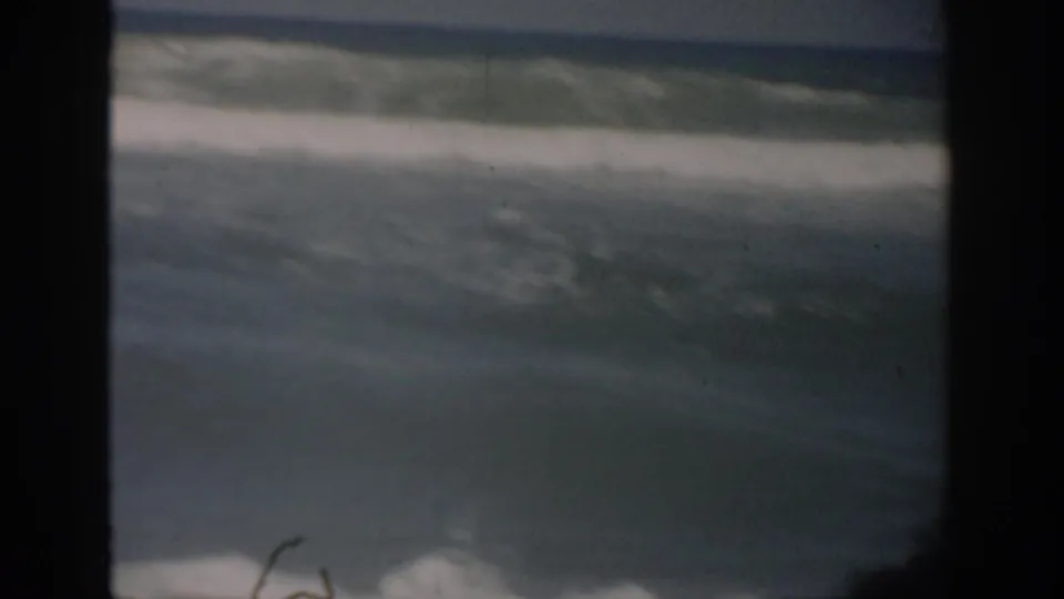1959 Florida 8mm home-movie frame showing ocean surf rolling toward shore under a pale sky, with dark film borders visible.