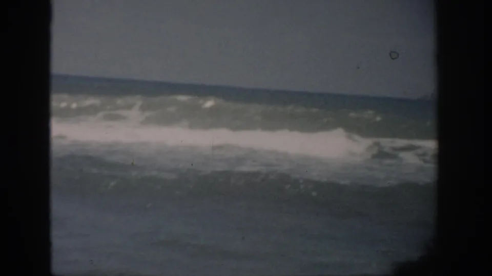 1959 Florida home-movie frame with ocean swell, whitewater, and a pale sky, showing muted color and 8mm gate borders.