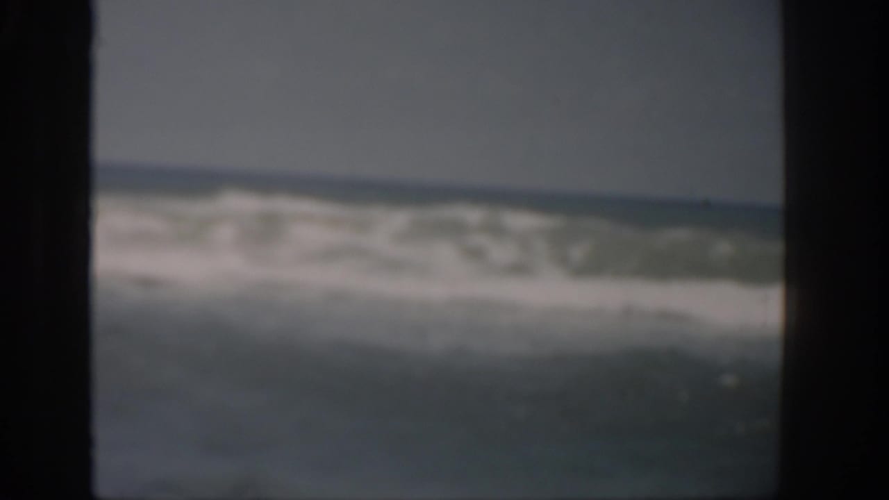 1959 Florida 8mm frame showing white surf and blue-gray ocean water approaching a sandy shoreline under a dull sky.