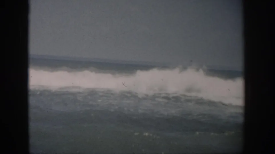 1959 Florida home-movie frame with a white breaker spreading across the ocean surface, seen through dark projected-frame edges.