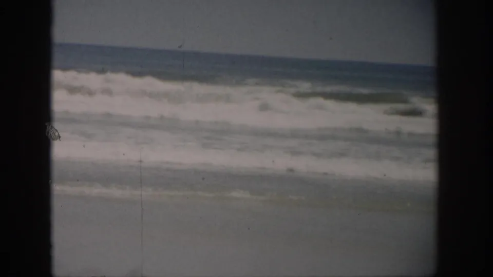 1959 Florida 8mm frame showing surf washing toward a sandy beach, bordered by dark film edges and softened by grain.