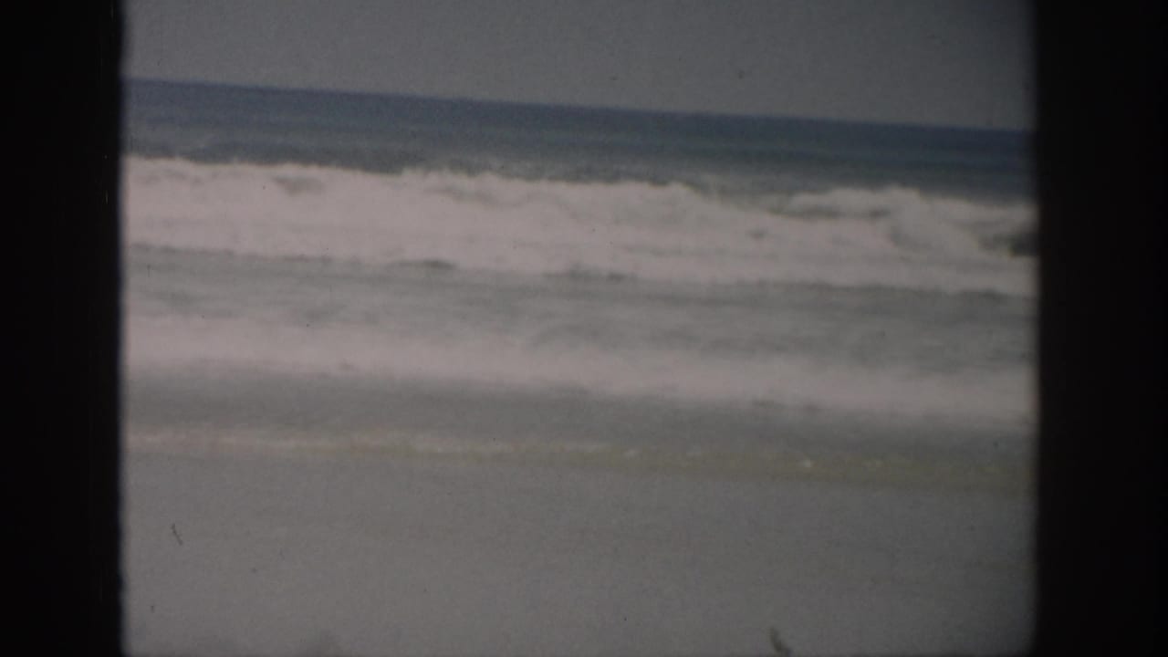 1959 Florida home-movie frame showing ocean waves breaking near a sandy beach, with dark film borders and faded color.