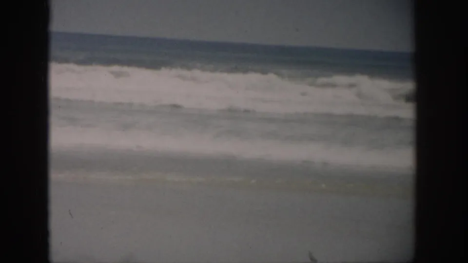 1959 Florida home-movie frame showing ocean waves breaking near a sandy beach, with dark film borders and faded color.