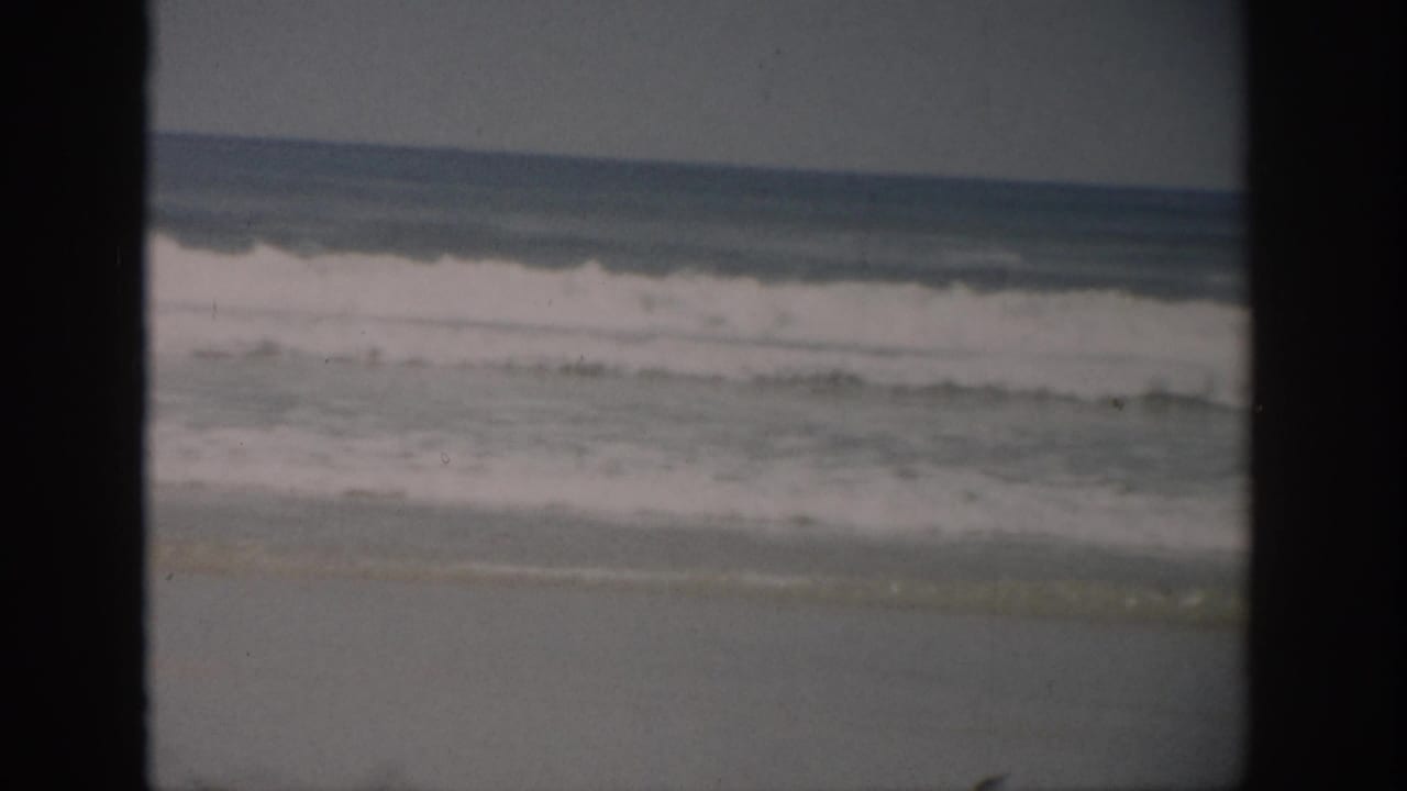 1959 Florida shoreline view with white-capped surf, wet sand, muted sky, and visible 8mm frame borders.