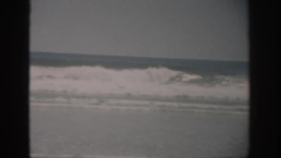 1959 Florida beach footage frame showing foamy waves moving shoreward, softened by faded 8mm color and dark frame edging.