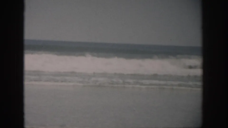 1959 Florida 8mm frame of a broad, quiet shoreline with white waves breaking across the middle distance.
