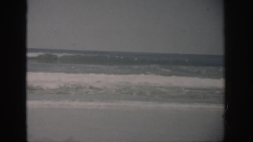 1959 Florida coastal frame with white surf bands, sandy beach, gray sky, and prominent dark borders from the 8mm scan.