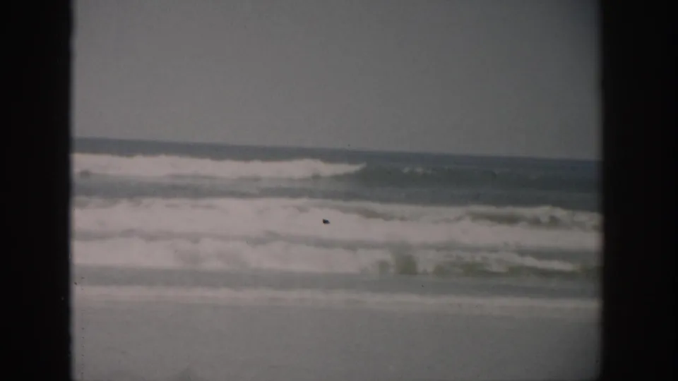 1959 Florida 8mm still of gentle ocean surf crossing a sandy beach, with faded color and visible home-movie grain.