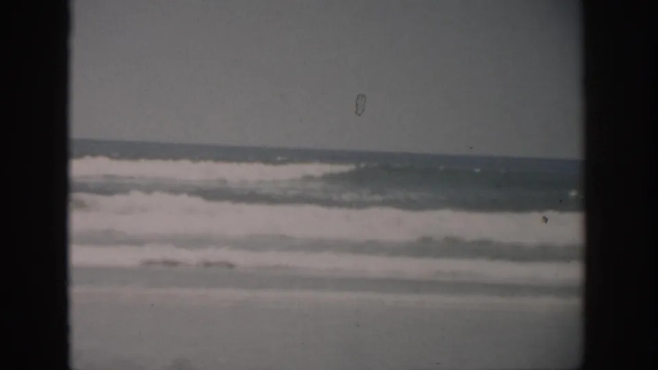 1959 Florida beach frame showing low ocean waves, a flat horizon, sandy shore, subdued color, and small marks from the 8mm film scan.