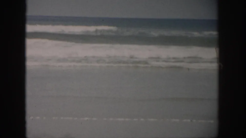 1959 Florida shoreline frame with layered surf, wet sand, muted color, and visible 8mm film borders from the scan.