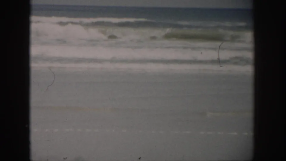 1959 Florida coastal frame with breaking waves, pale foam, wet beach, and visible film scratches near the frame edge.