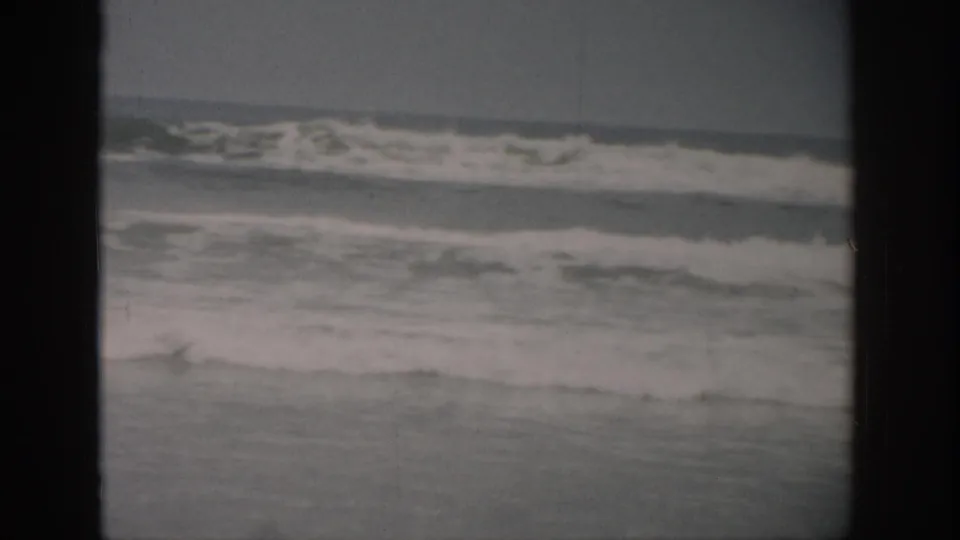 1959 Florida beach frame showing choppy surf angled across the shoreline, with muted sky and archival film grain.
