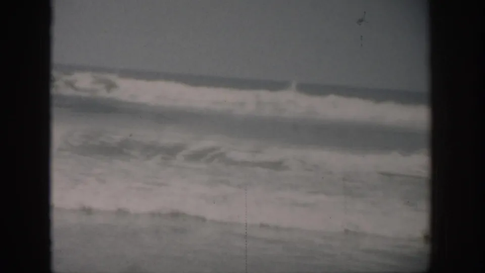 1959 Florida 8mm frame of foamy ocean surf rolling in under an overcast sky, with visible film scratches and gate borders.