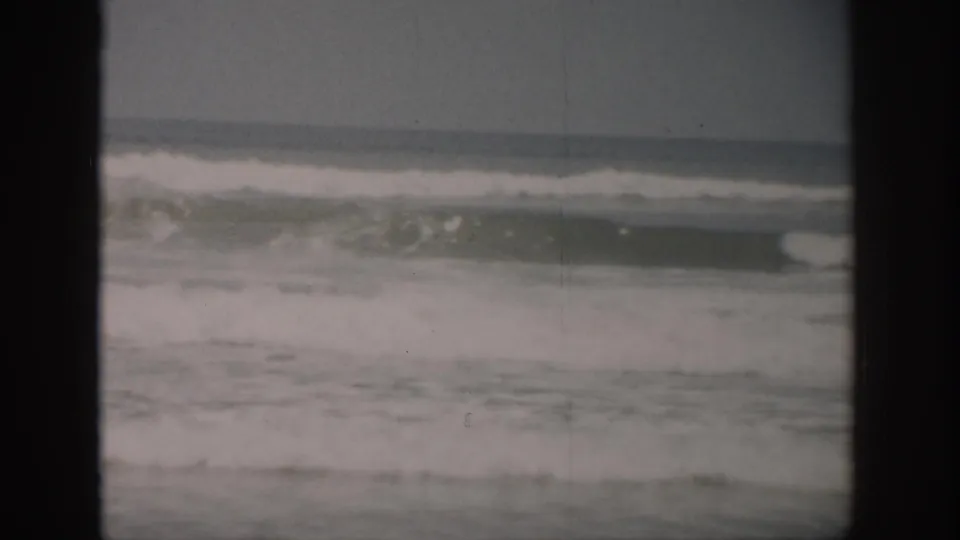 1959 Florida shoreline frame showing foamy surf under a gray sky, recorded with visible 8mm grain, dust, and gate framing.