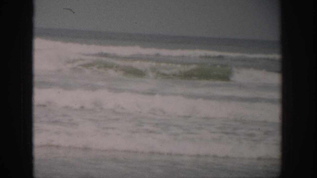 1959 Florida home-movie frame showing low ocean surf, white foam, wet sand, and an overcast horizon.