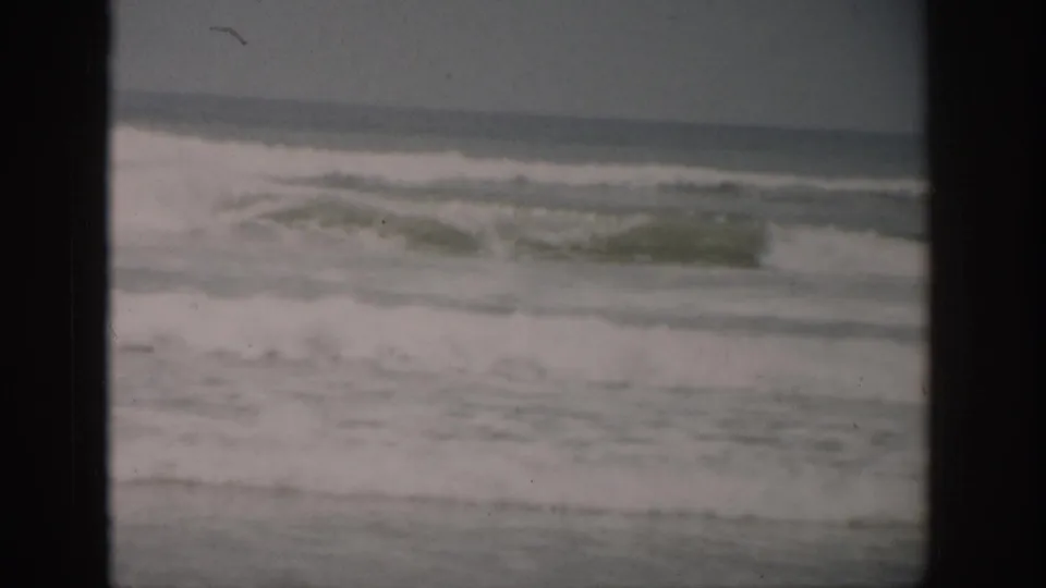 1959 Florida home-movie frame showing low ocean surf, white foam, wet sand, and an overcast horizon.