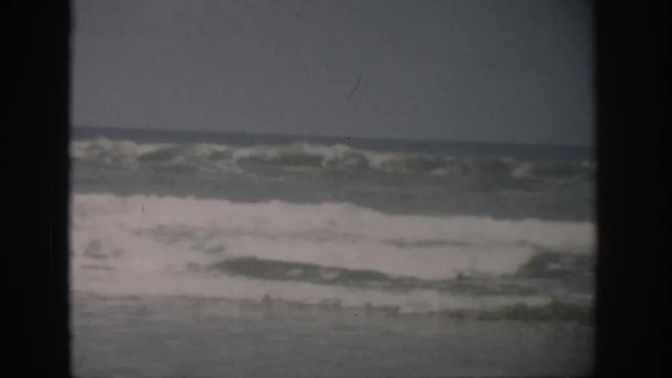 Florida coastal home-movie frame from 1959 showing foamy waves moving over wet sand beneath a dark horizon line.