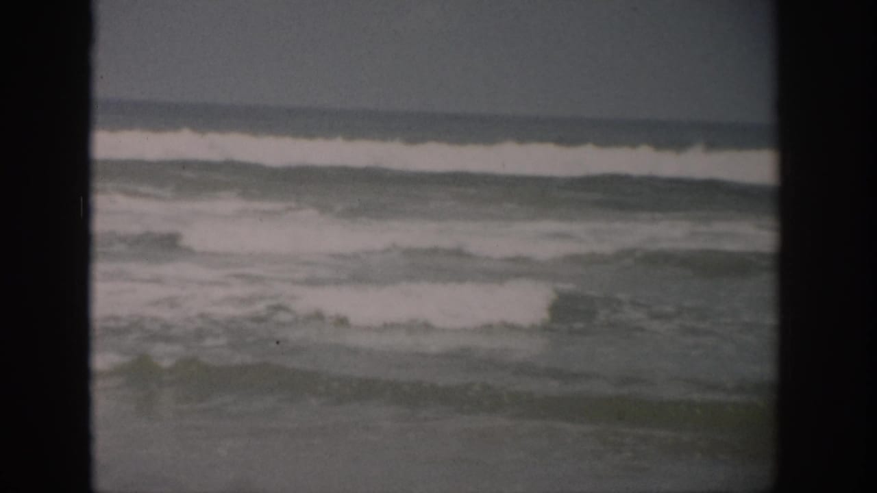 Small surf rolls toward shore beneath a muted gray sky, framed by dark film borders.